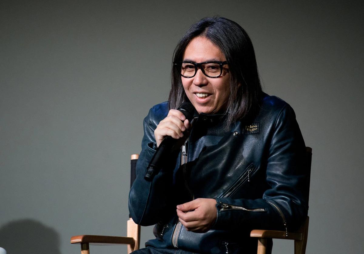 A man with long hair and glasses in a black leather jacket seated on stage speaking into a handheld microphone against a plain grey backdrop.