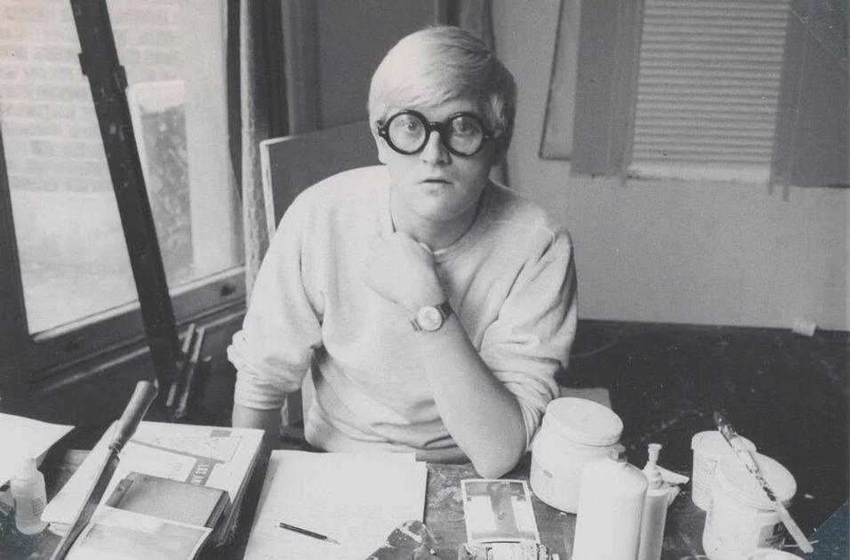 A black and white photograph of David Hockney as a young man in thick round glasses and a pale sweater, seated at a work table crowded with paint pots, tubes and brushes.