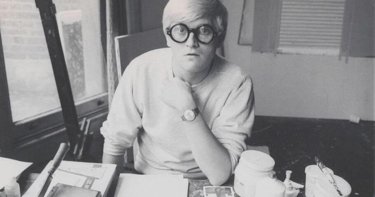 A black and white photograph of David Hockney as a young man in thick round glasses and a pale sweater, seated at a work table crowded with paint pots, tubes and brushes.