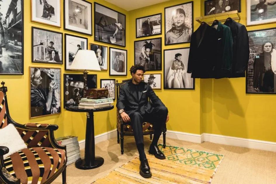 A man in a dark suit seated on a wooden chair in a yellow walled room hung floor to ceiling with framed black and white photographs, coats on a rail beside him and a striped rug underfoot.