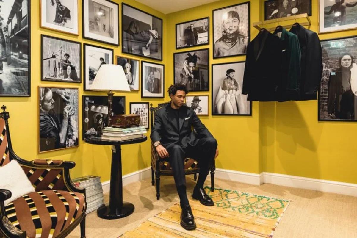 A man in a dark suit seated in a yellow walled room hung floor to ceiling with framed black and white photographs, coats on a rail beside him.