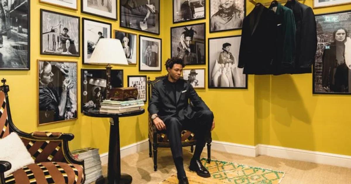 A man in a dark suit seated on a wooden chair in a yellow walled room hung floor to ceiling with framed black and white photographs, coats on a rail beside him and a striped rug underfoot.