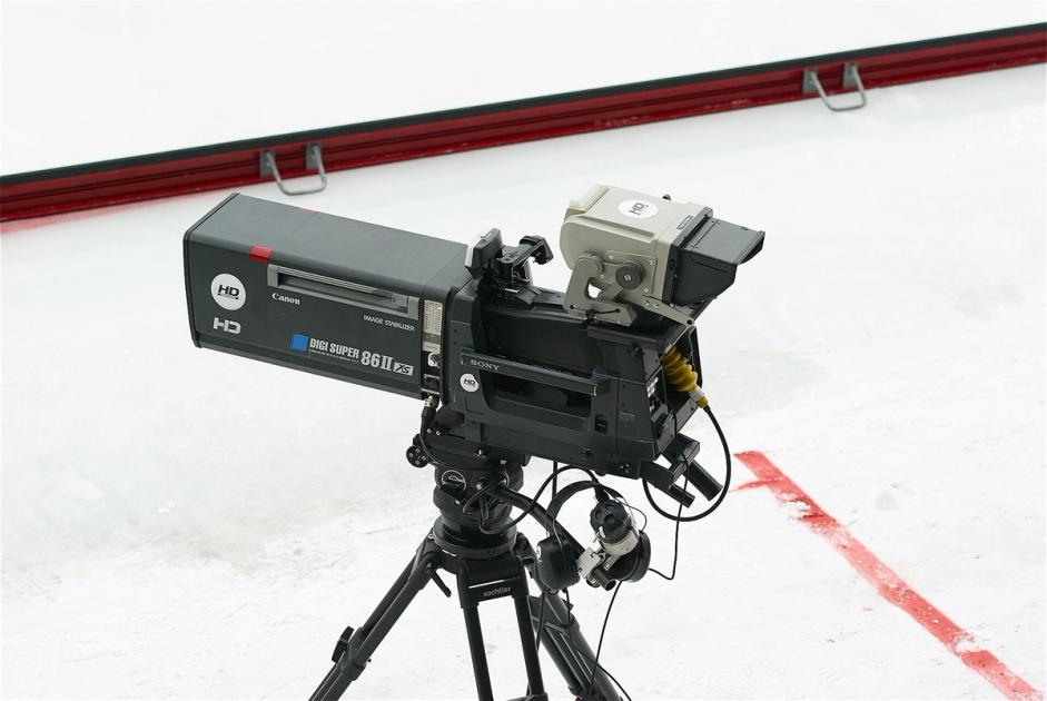 A professional broadcast television camera with a long lens, mounted on a tripod beside the boards of an ice rink.