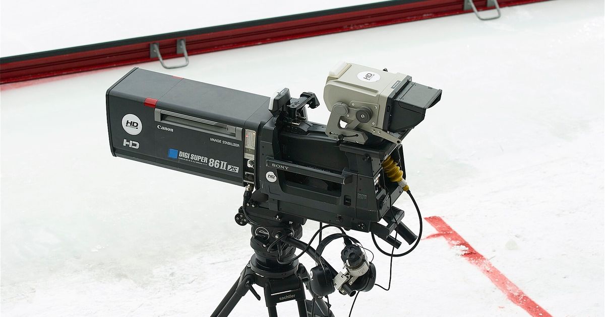 A professional broadcast television camera with a long lens, mounted on a tripod beside the boards of an ice rink.