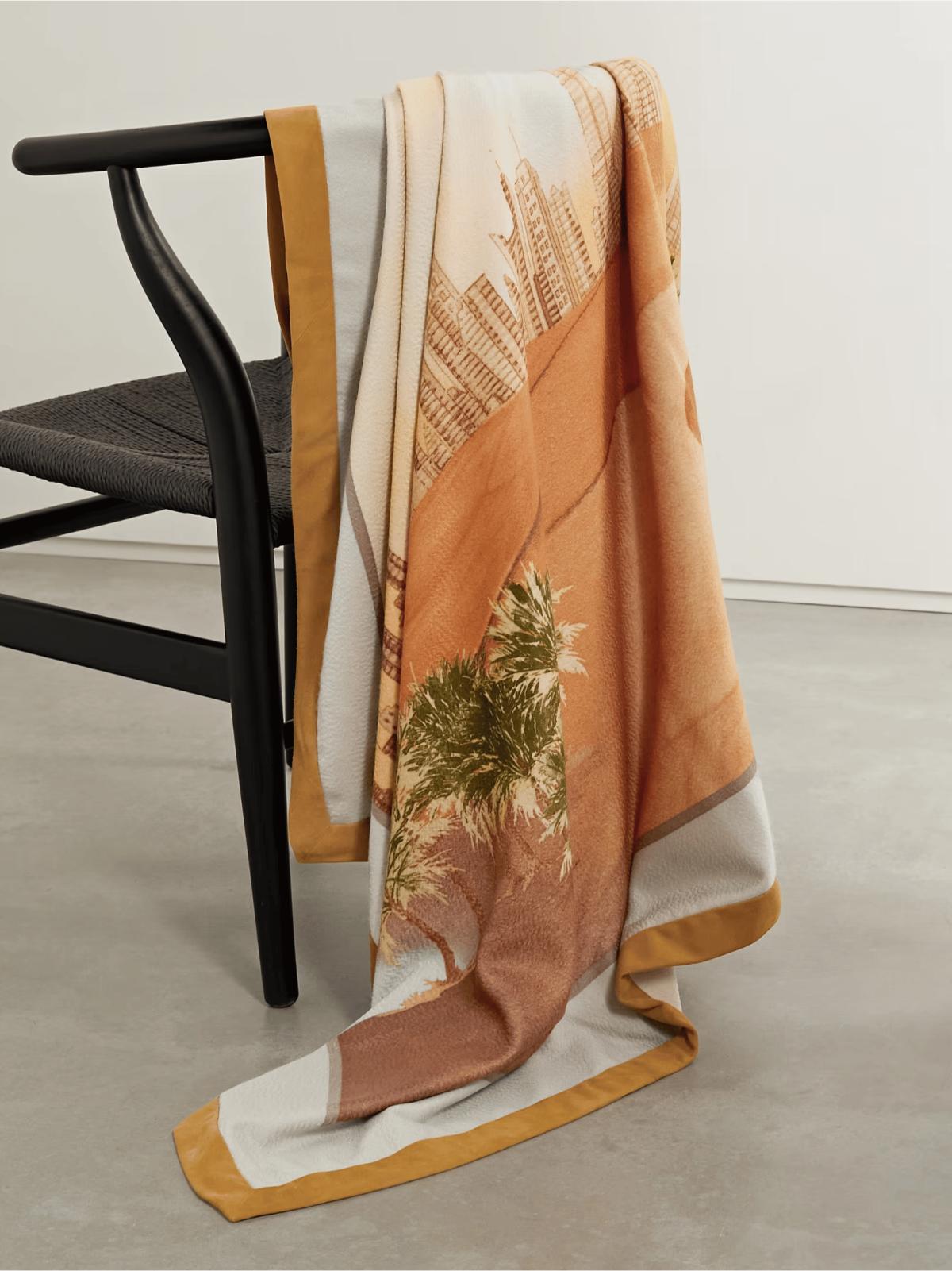A large printed silk scarf in apricot, cream and gold draped over the back of a black wishbone chair against a pale wall.