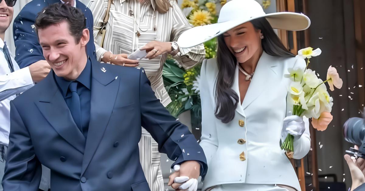 A couple leaving a building through thrown confetti, he in a navy double breasted suit, she in a white trouser suit and wide brimmed hat holding a bunch of narcissus.