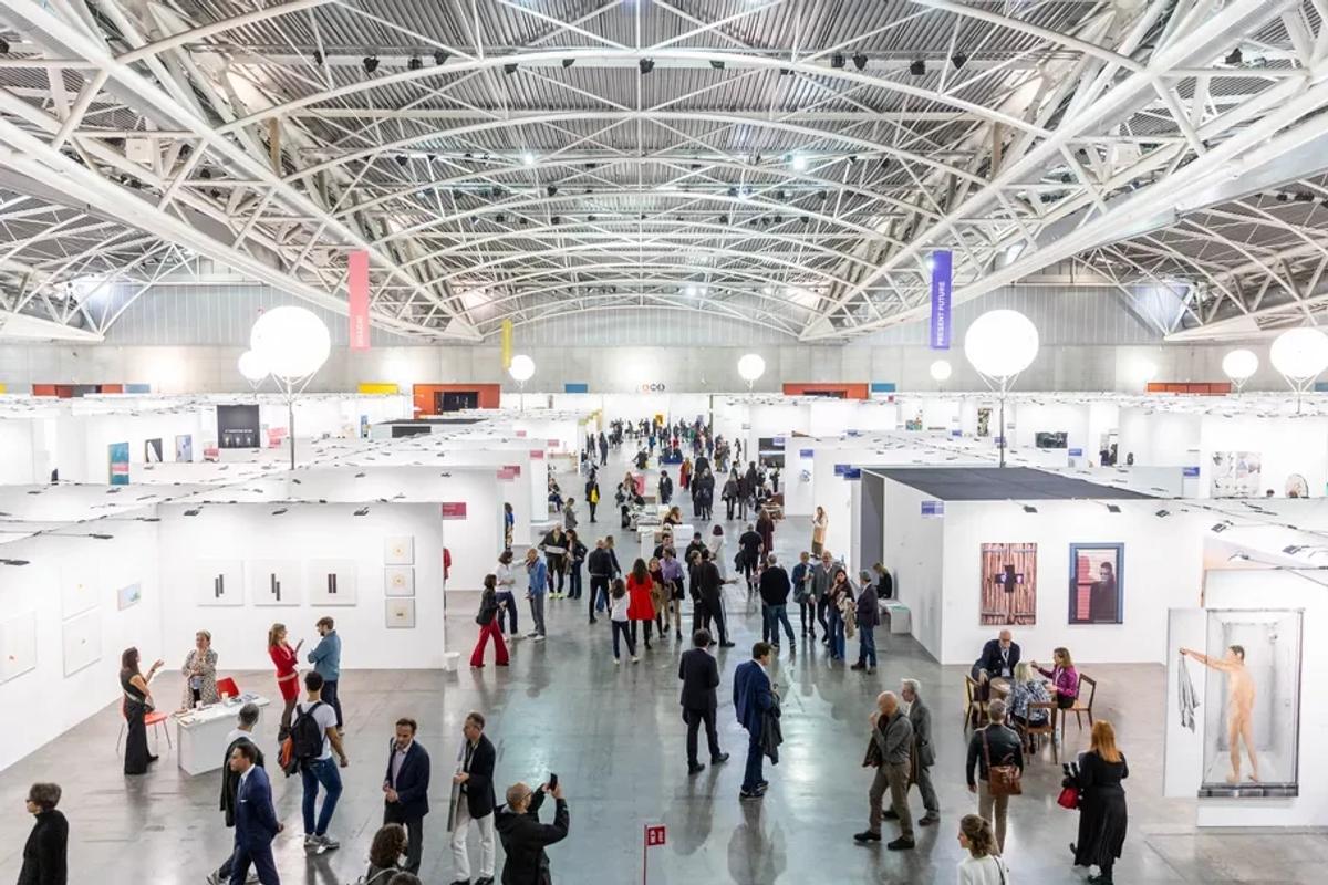 An art fair hall seen from above under a white lattice roof, rows of white booths and crowds of visitors moving between them.