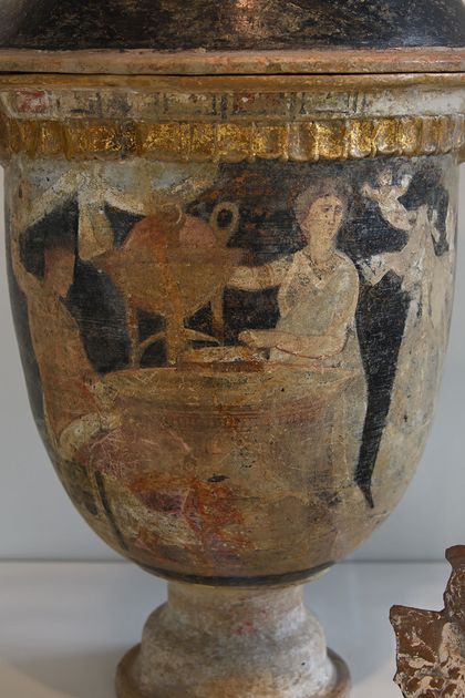 A tall footed ceramic vase with a gilded moulded rim, painted with standing figures at an altar in muted reds and browns on a pale ground.