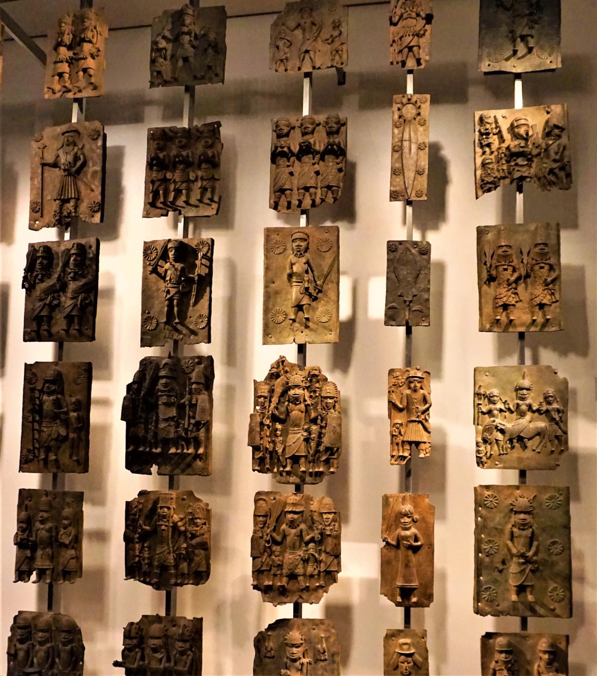 Benin bronze plaques displayed in a glass case at the British Museum, cast brass reliefs showing figures in ceremonial dress.
