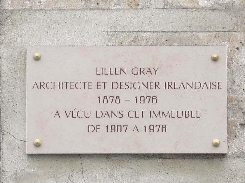 A pale stone plaque fixed to a Paris wall, reading EILEEN GRAY, ARCHITECTE ET DESIGNER IRLANDAISE, 1878 to 1976, lived in this building from 1907 to 1976.