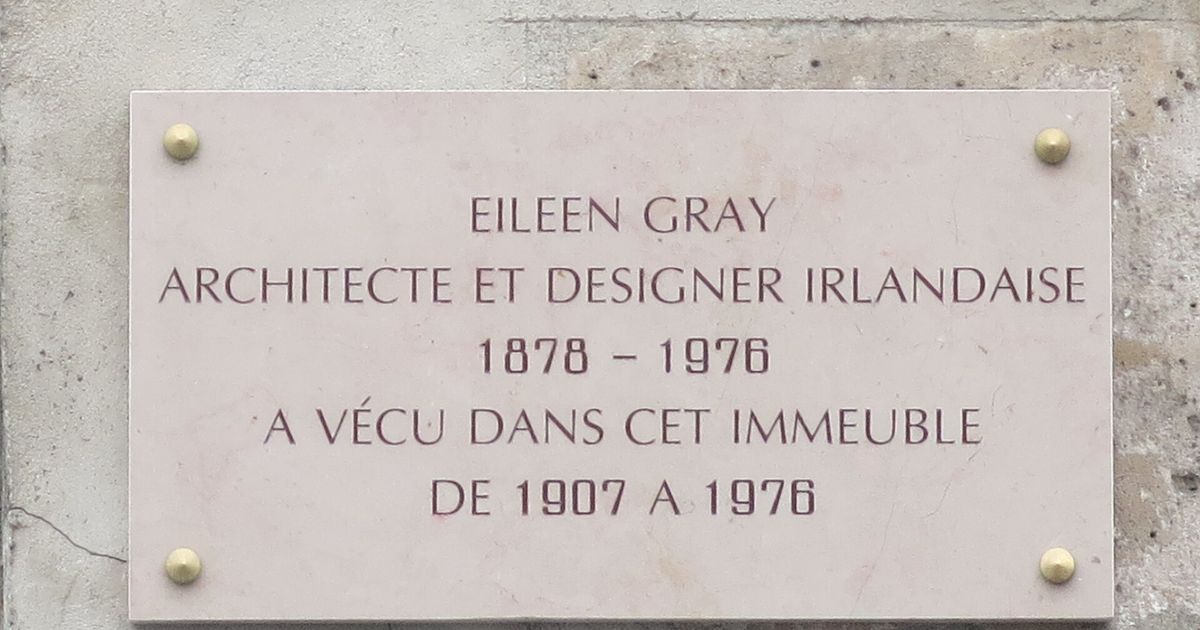 A pale stone plaque fixed to a Paris wall, reading EILEEN GRAY, ARCHITECTE ET DESIGNER IRLANDAISE, 1878 to 1976, lived in this building from 1907 to 1976.