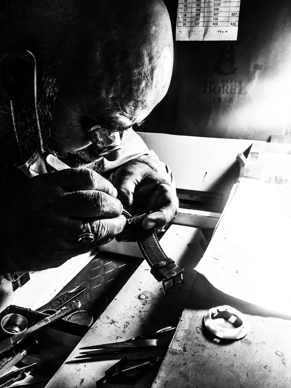 A watchmaker at a bench working on a wristwatch movement with tweezers, parts and tools laid out around it.
