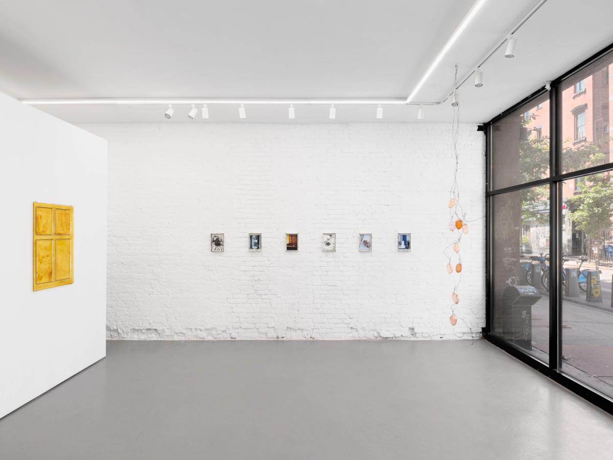 Wide gallery view: an amber glass window panel on a white wall at left, six small mirrored-glass photographs along a white brick wall at centre, and the glass lemons of Lemon Tree hanging by the storefront window at right.
