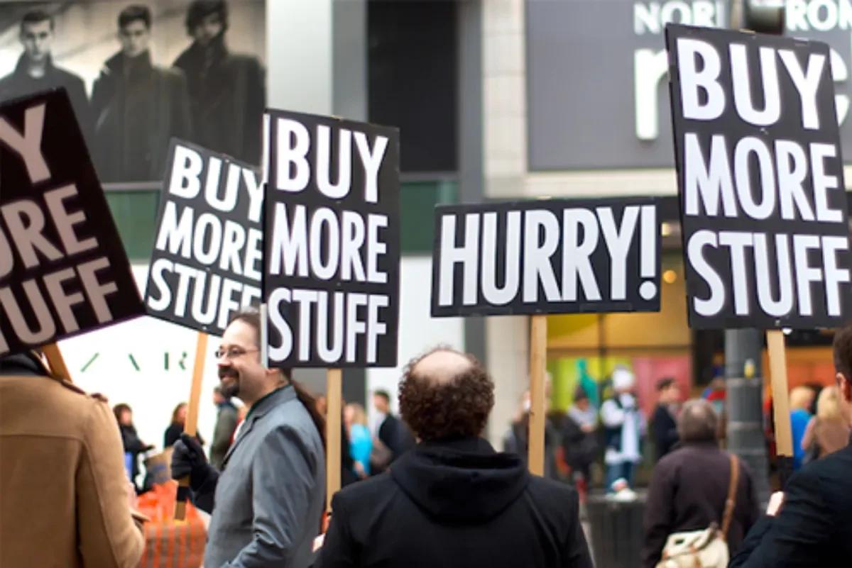 A crowd in a street holding placards printed with the words buy more stuff and hurry, photographed from behind.