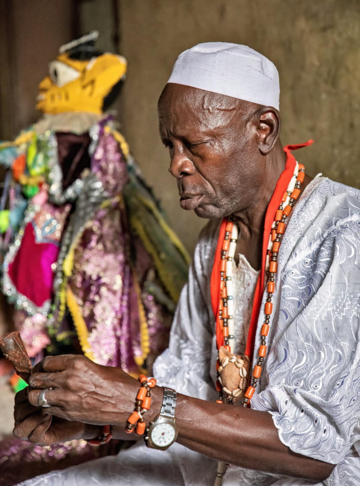 An older man in a white cap and embroidered robe wearing layered orange bead necklaces, holding a beaded and sequinned figure beside him.