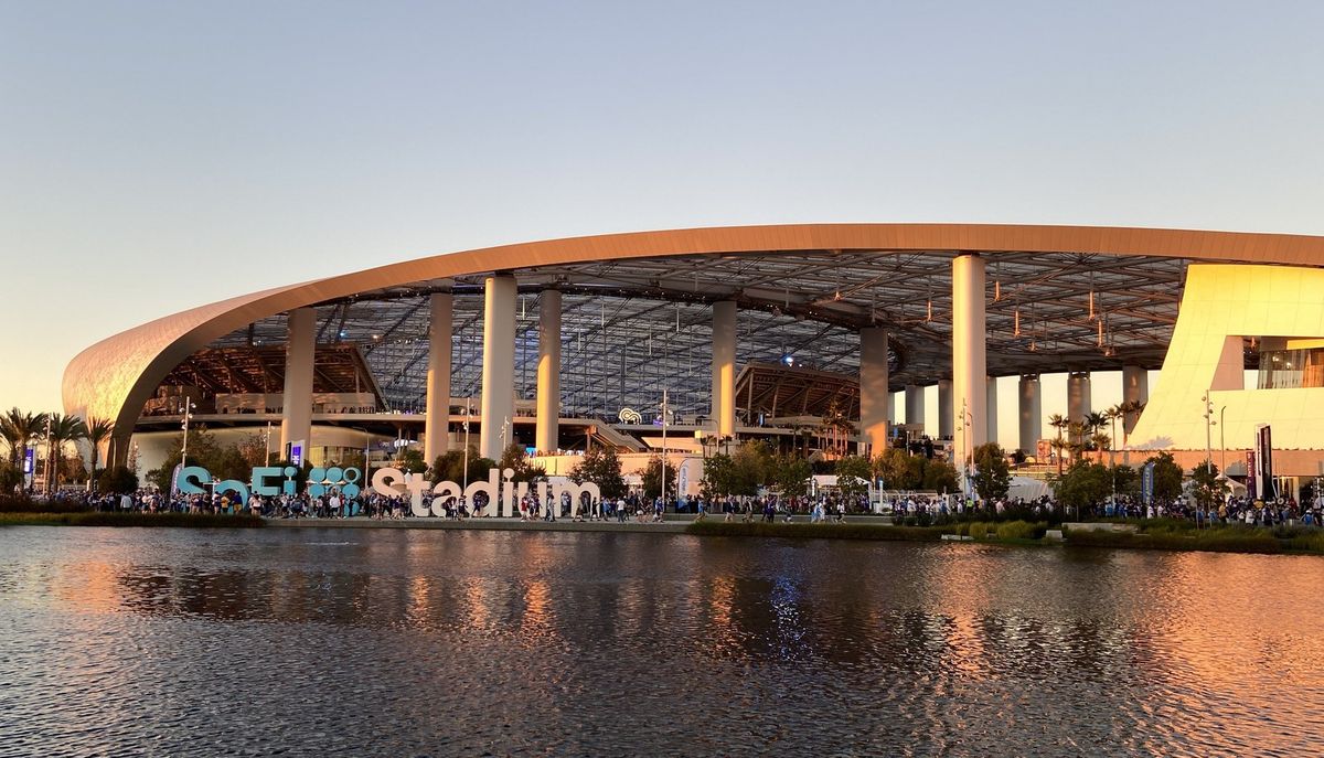 The exterior of SoFi Stadium at dusk, its curved translucent canopy lit from beneath, the words SoFi Stadium standing in large letters beside a lake, with spectators walking along the water in front of it.