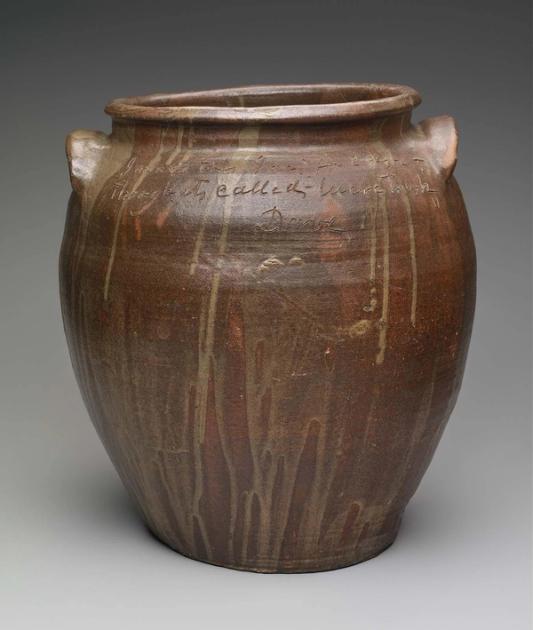 A large brown alkaline-glazed stoneware storage jar with two lug handles, drips of lighter glaze running down its sides, and a line of handwriting incised around the shoulder ending in the name Dave.