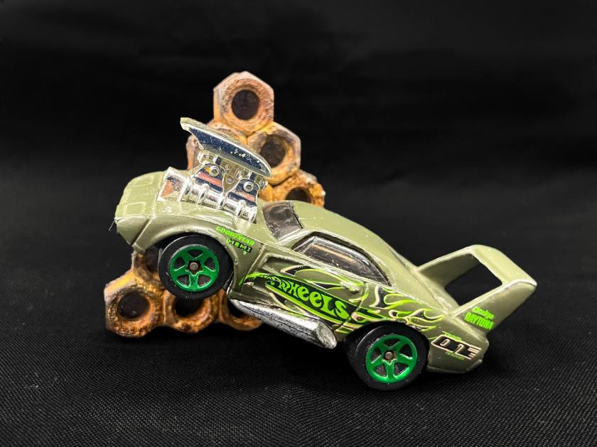 A brooch made from a Hot Wheels toy car mounted on a pale ground, with a stack of weathered steel nuts soldered to its rear deck.