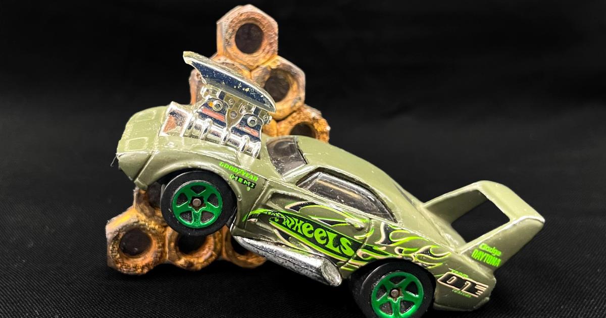 A brooch made from a Hot Wheels toy car mounted on a pale ground, with a stack of weathered steel nuts soldered to its rear deck.