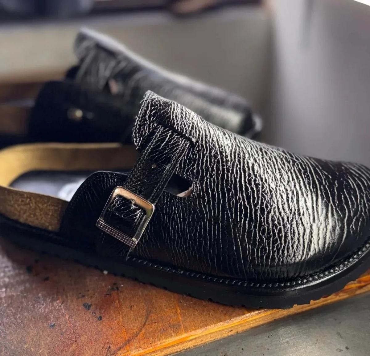 A buckled mule in heavily grained black leather on a cork footbed, photographed close on a wooden bench.