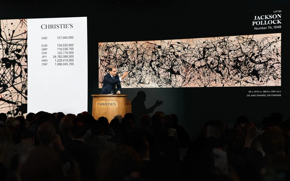 An auction room at Christie's, the auctioneer at a lit rostrum before a wide screen showing a Jackson Pollock drip painting and a column of bids in several currencies, an audience seated in darkness.