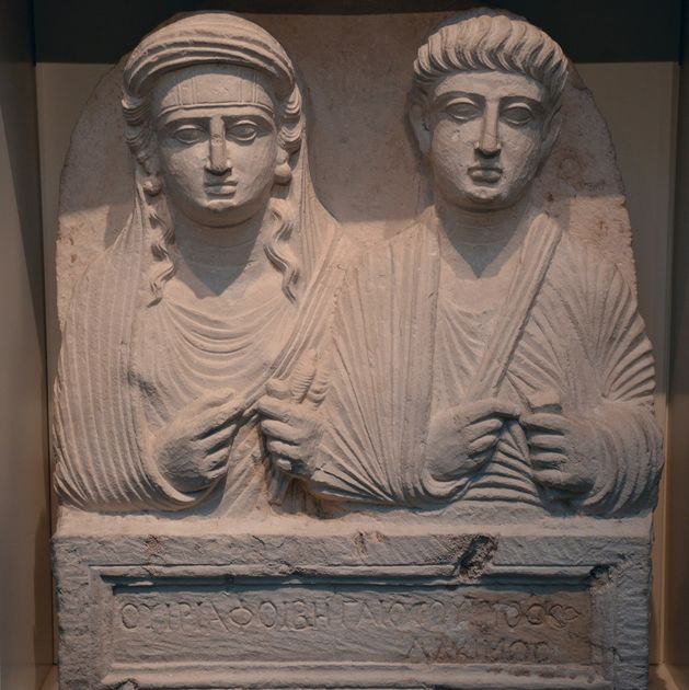 A carved limestone funerary relief from Palmyra showing the busts of a man and a veiled woman side by side, with an inscription cut beside them.