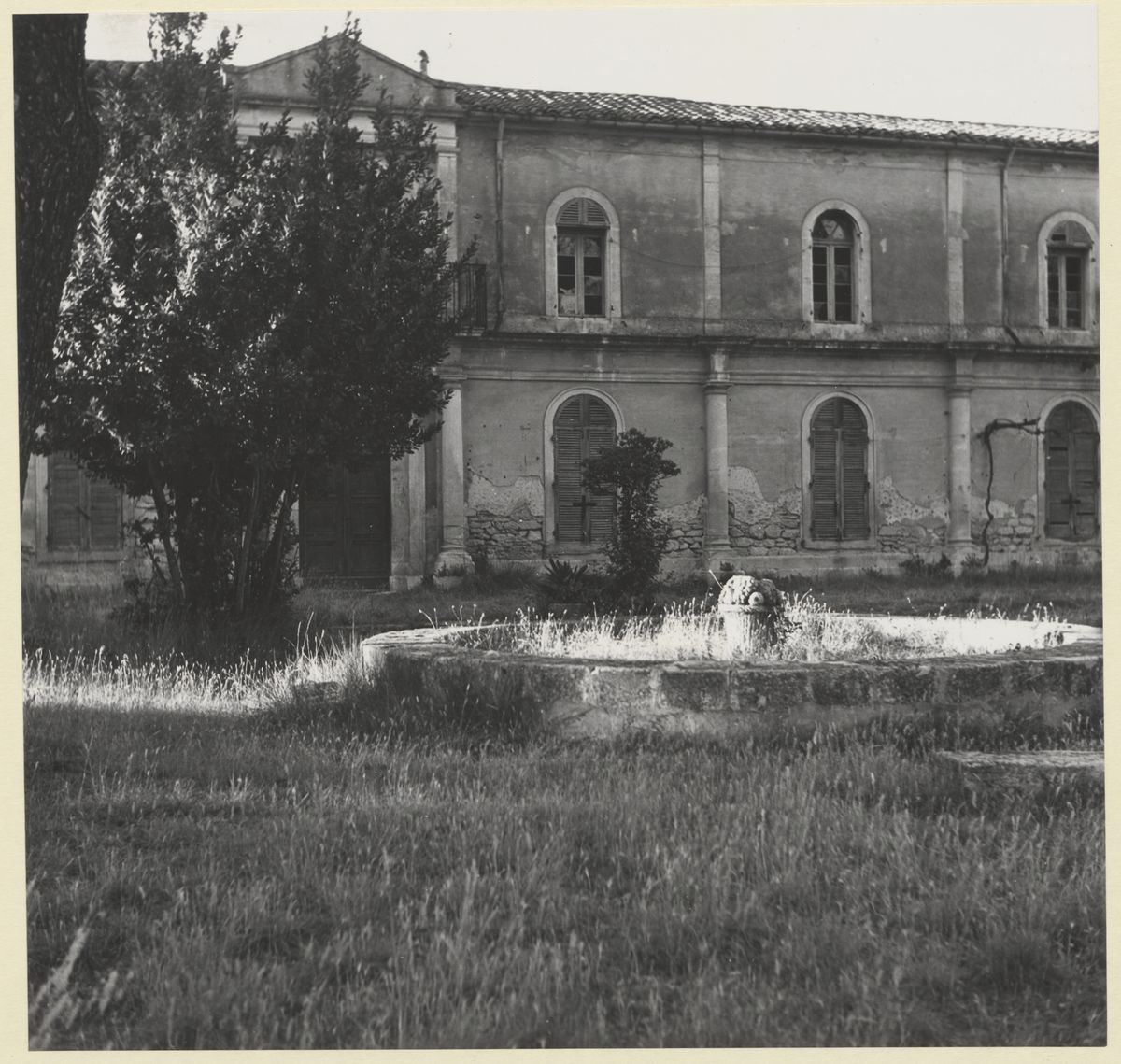 A stone monastery building behind a low wall, with an old fountain in the foreground and trees around it.