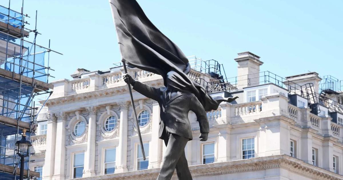 A sculpture of a suited figure whose head is replaced by a large black flag, mounted high against a pale stone building and blue sky.