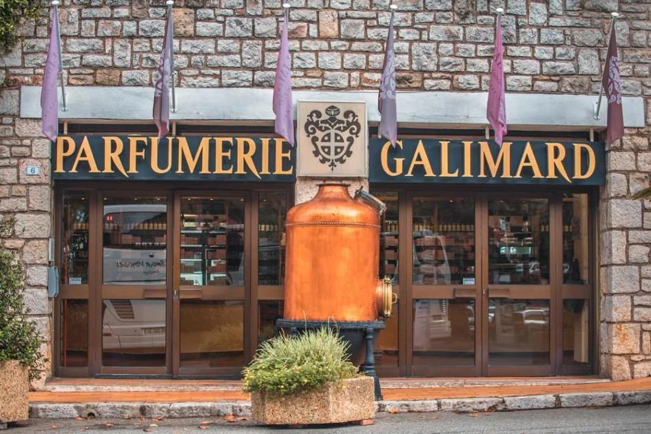 A stone fronted shop with Parfumerie Galimard in gold lettering above the windows, lavender coloured banners along the facade and a large copper still standing outside the door.