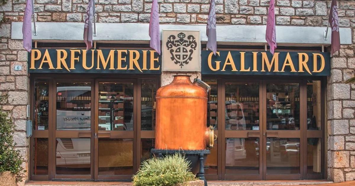 A stone fronted shop with Parfumerie Galimard in gold lettering above the windows, lavender coloured banners along the facade and a large copper still standing outside the door.