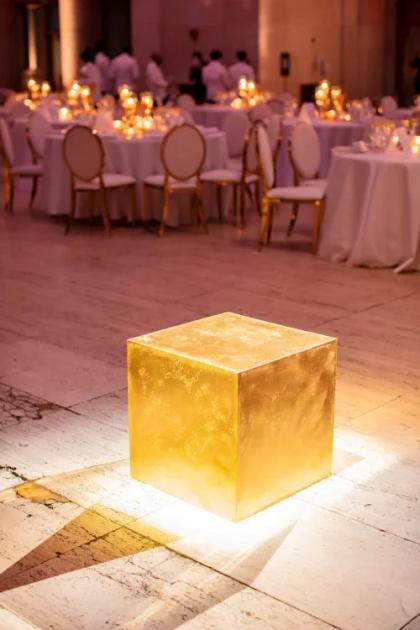A solid gold cube lit on a stone floor at a formal dinner, with candlelit round tables, gilt chairs and standing guests blurred behind it.
