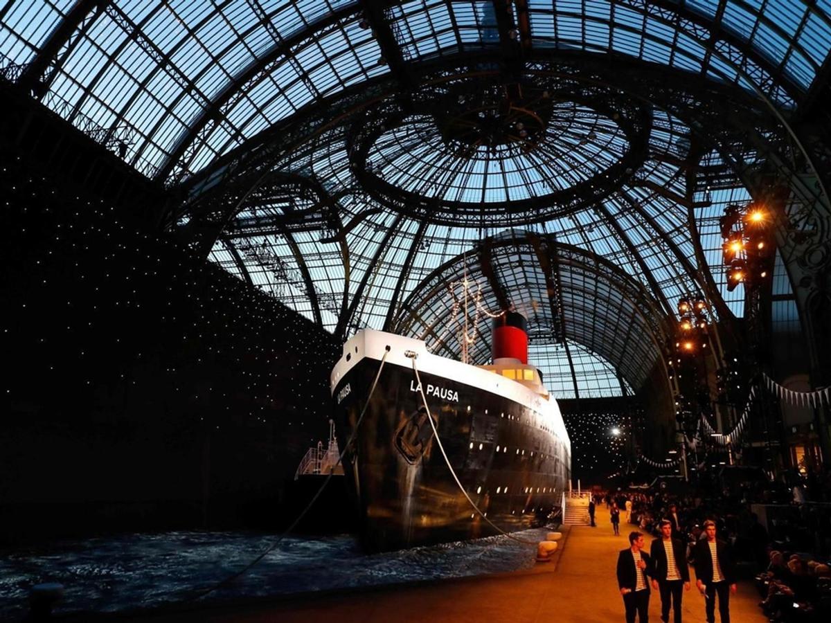 The Chanel Cruise 2019 show, featuring the colossal 'La Pausa' ship replica erected inside the Grand Palais. This is direct visual evidence that inter-seasonal collections are prioritized not as artisanal output, but as major global PR events and expensive marketing collateral designed purely to generate essential media buzz and continuous consumer engagement.