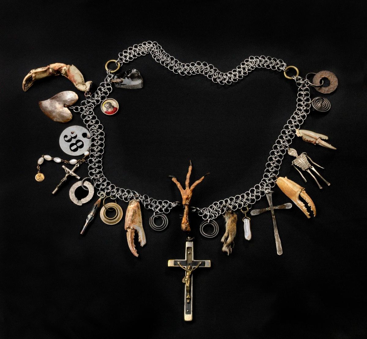 A chain necklace hung with found objects, including small cast hands, discs, keys and a crucifix, laid out against black.
