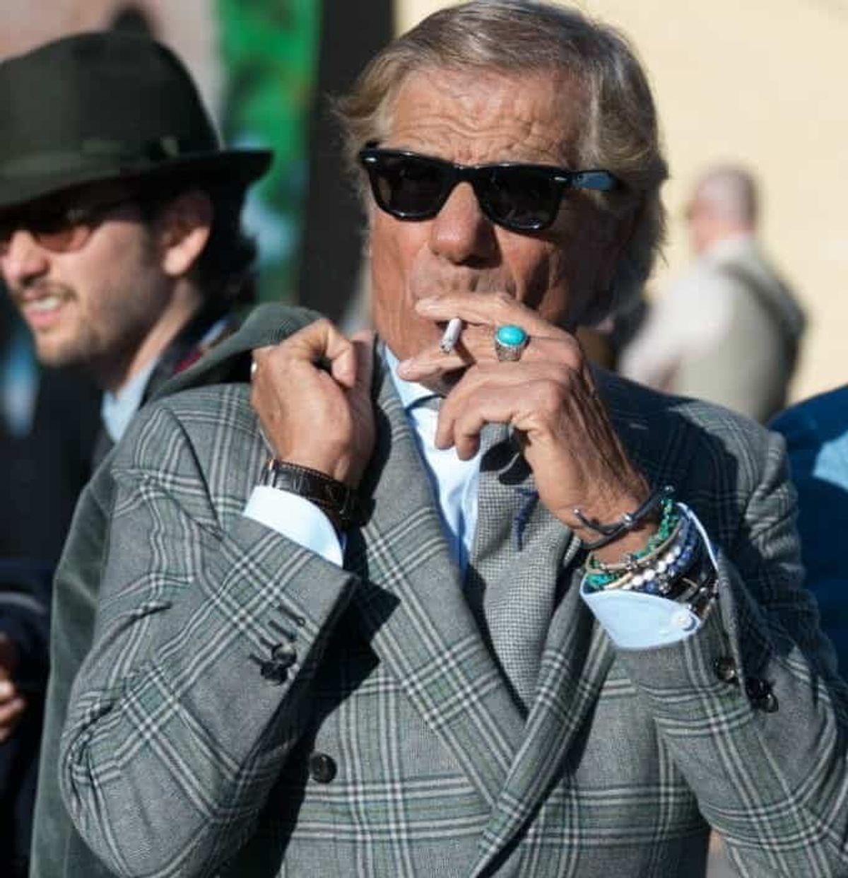 Lino Ieluzzi, the epitome of Italian style, captured in his element. A jacket rests perfectly on his shoulders, not worn but draped. His shirt cuffs are artfully unfastened, revealing a collection of wristbands, while a statement ring and classic sunglasses complete the look. The cigarette held with practiced ease is the final touch in this masterclass in sprezzatura