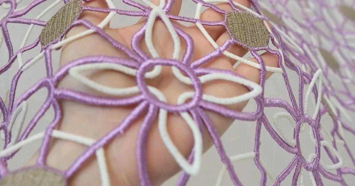 A close view of an open lace structure of lilac and cream cords forming linked flower shapes, held over skin.