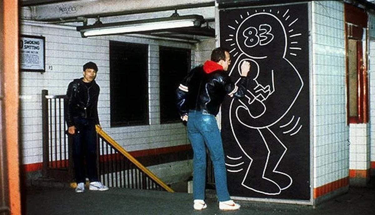 The genesis of a vocabulary before its financialization: Keith Haring executing a chalk drawing in the New York City subway system in 1983. This raw, ephemeral street intervention represents a pure anti-market ethos; a structural contrast to the 2026 Cruise show, where the corporate apparatus archives and sanitizes this exact downtown subversion to manufacture luxury legitimacy.