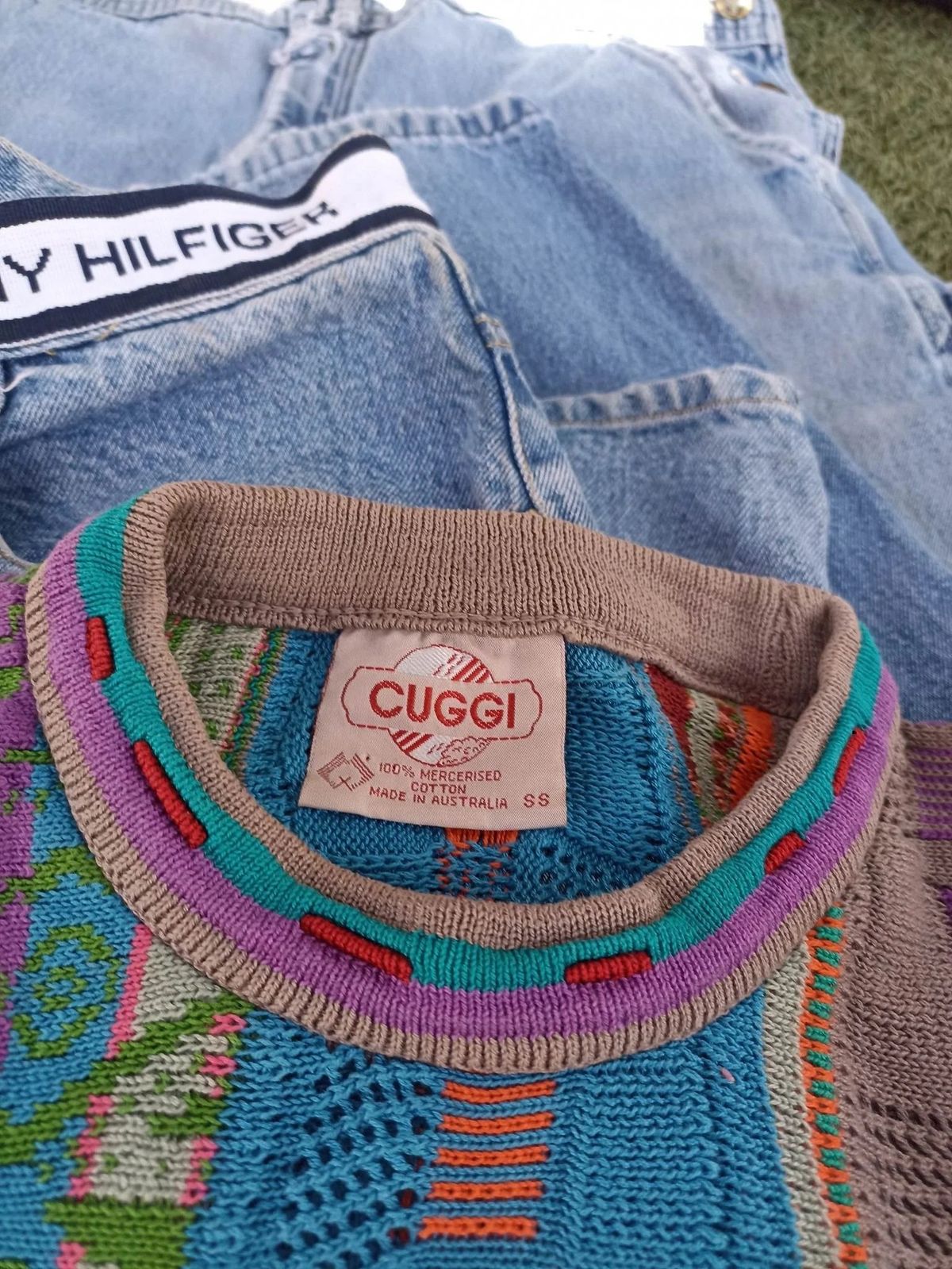 A folded pair of pale jeans with a branded waistband, laid over a knitted sweater in blue, pink and green whose neck label reads Cuggi.