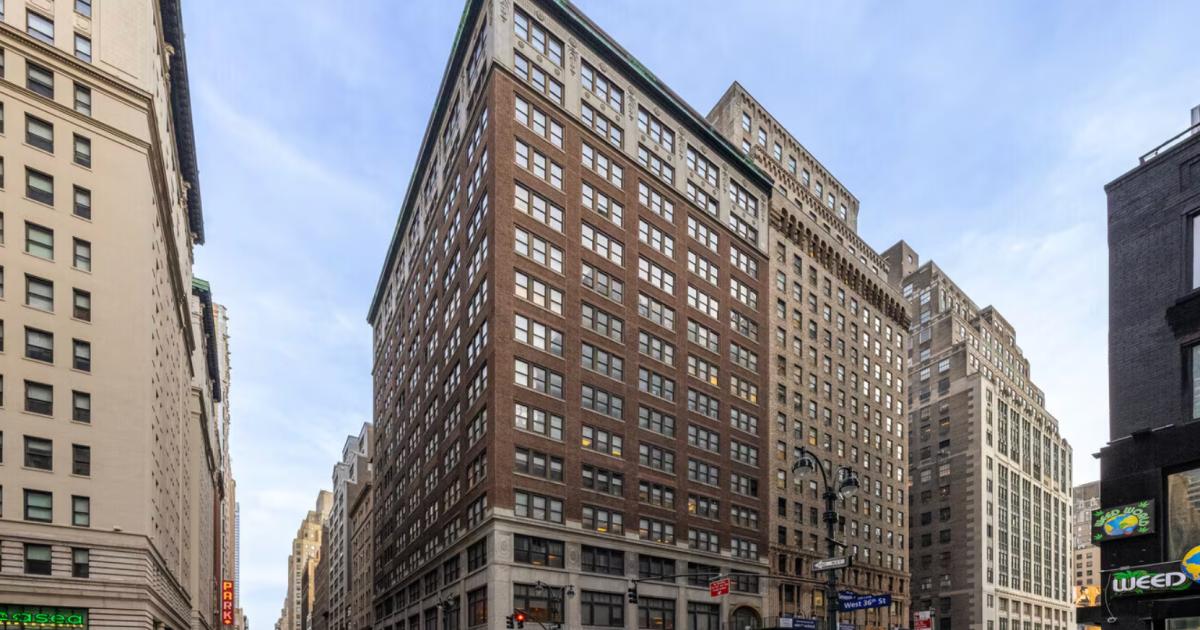 A tall brick and limestone office building on a Manhattan corner under a blue sky, older towers either side, traffic and shopfronts at street level.