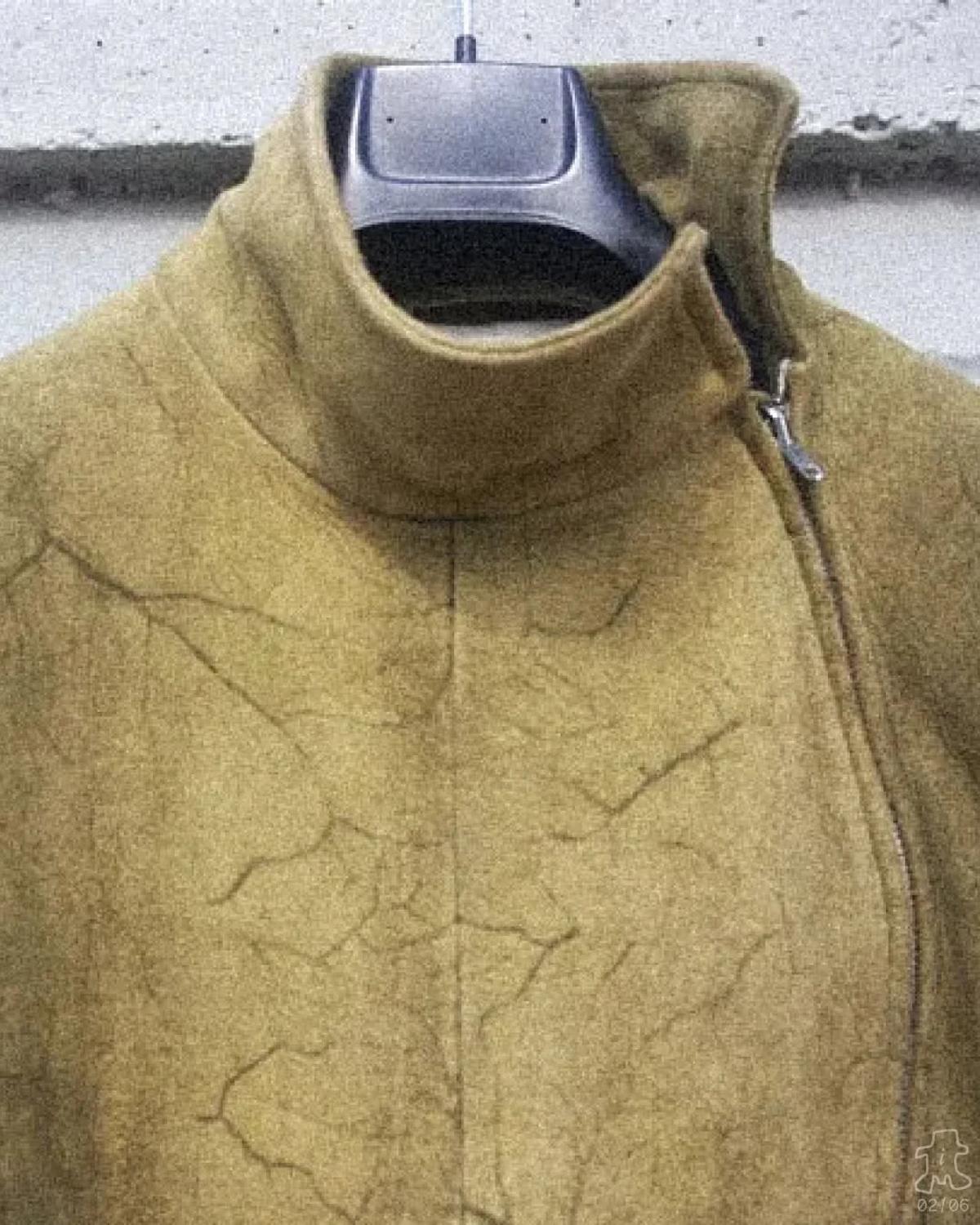 The upper half of an olive coated jacket on a hanger, its high funnel collar and chest showing a fine cracked surface like dried mud.