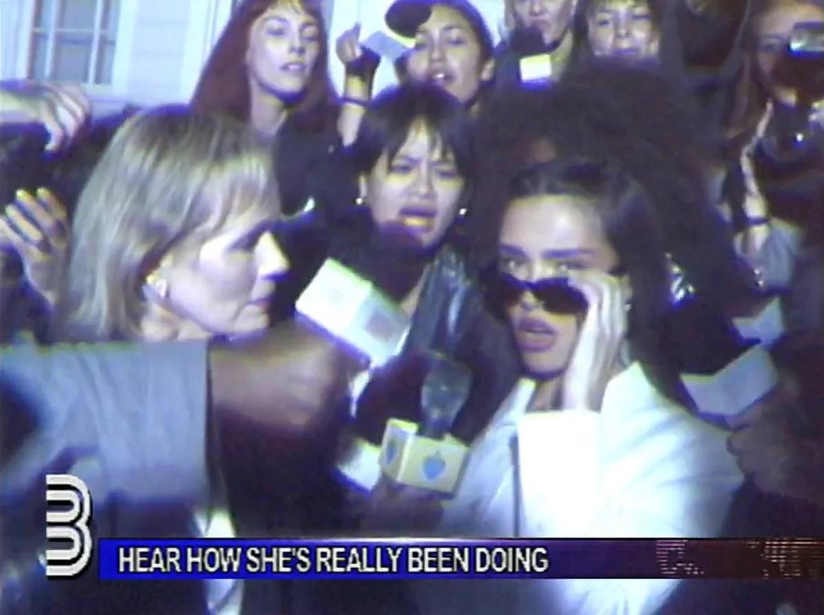 A television news still of a woman in dark glasses pressed by a crowd of reporters holding microphones, a caption bar across the lower edge.