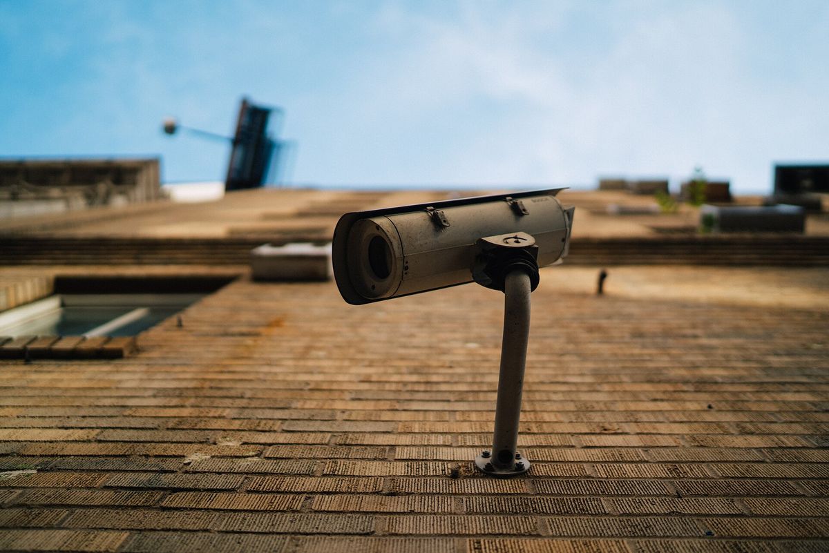 A single street surveillance camera mounted on a brick wall, photographed from below against a pale sky. No people are in the frame.