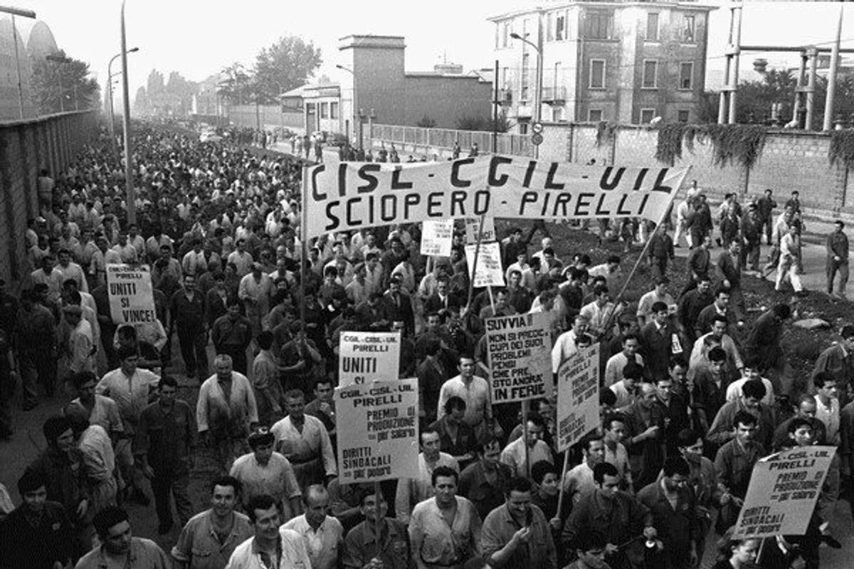 A black and white photograph of a large crowd of workers filling a street, carrying union banners and printed placards.