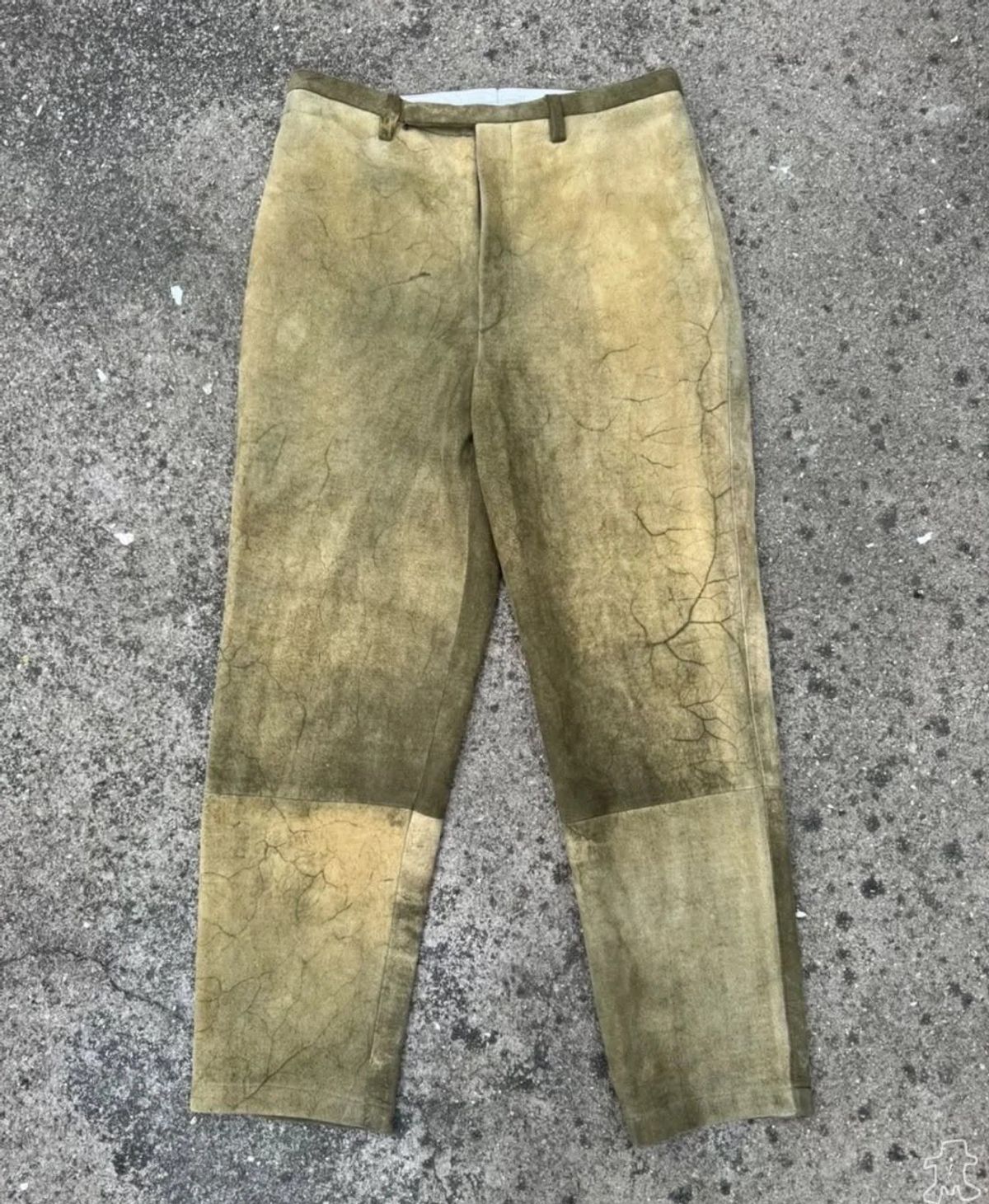 A pair of trousers in cracked olive coated fabric laid flat on rough concrete, the finish worn pale at the knees.
