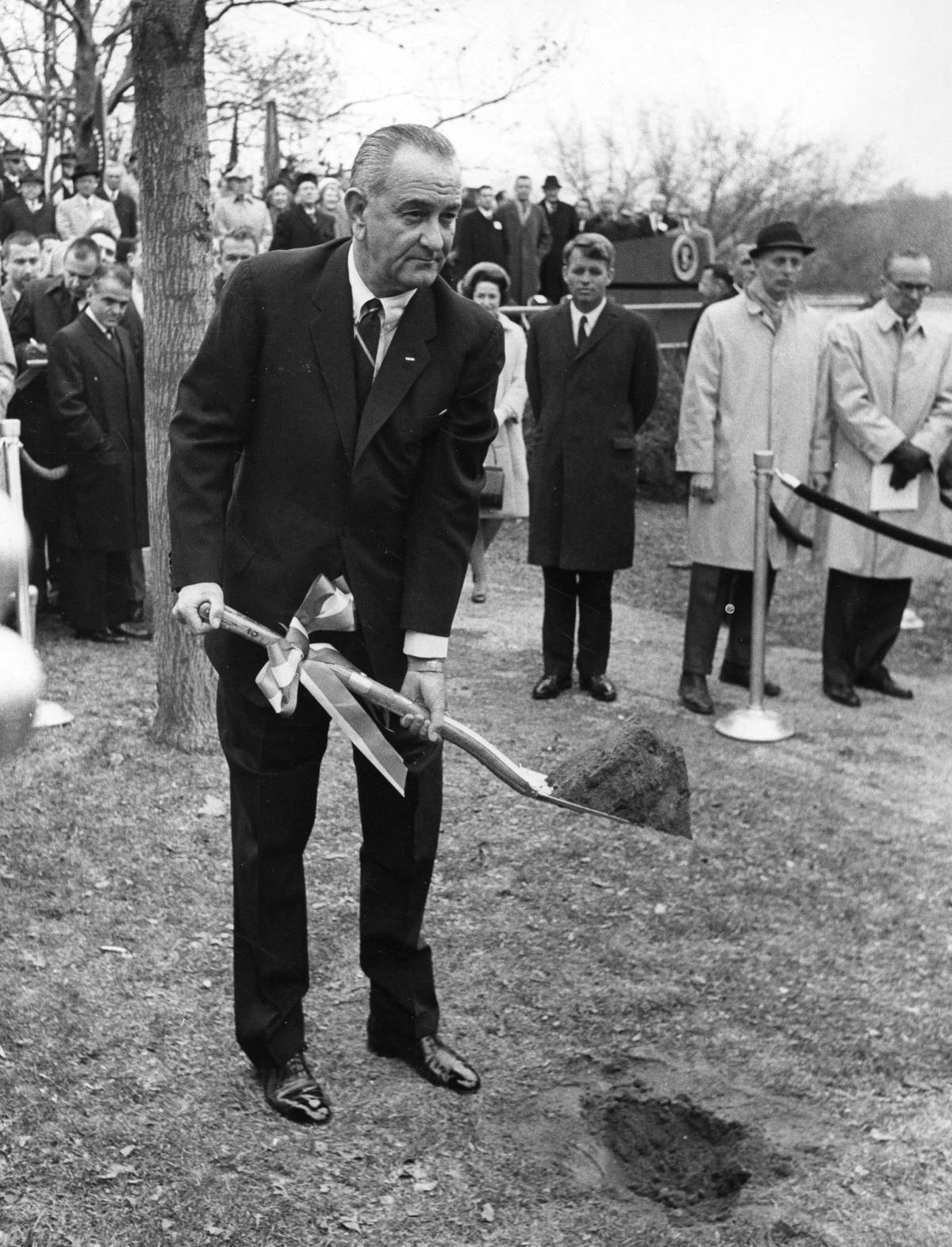Depositing the foundation of Labor Density: President Lyndon B. Johnson breaks ground on the Potomac shore on December 2, 1964, flanked by Senator-Elect Robert F. Kennedy. This material act established the building not as a real estate asset, but as a Sovereign Object designed to institutionalize national grief and secure Narrative Permanence through continuous custodial stewardship.