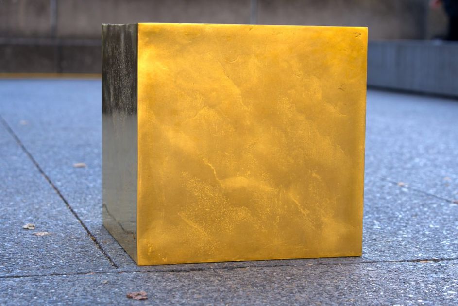 A gold surfaced cube standing on a paved city square, its side catching the light against grey stone slabs.