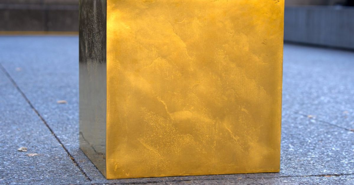 A gold surfaced cube standing on a paved city square, its side catching the light against grey stone slabs.