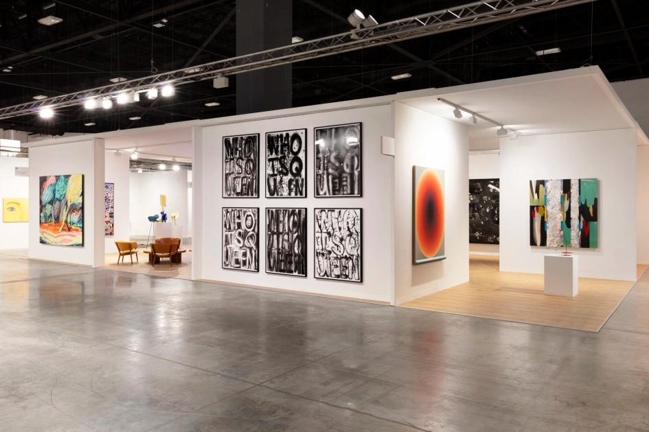 An art fair booth with white walls: six black and white text paintings hung in a grid, a red and black concentric canvas beside them, further works around a polished concrete floor.