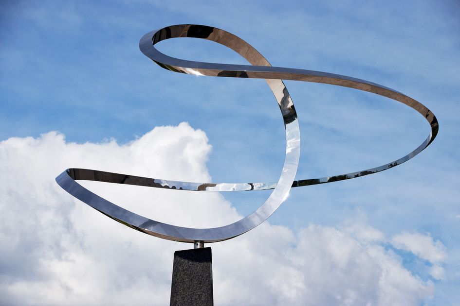 Infinity, a polished stainless steel ribbon sculpture on a dark granite pedestal, photographed against a sky of blue and white cloud.