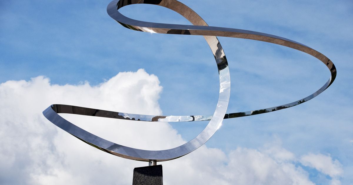Infinity, a polished stainless steel ribbon sculpture on a dark granite pedestal, photographed against a sky of blue and white cloud.