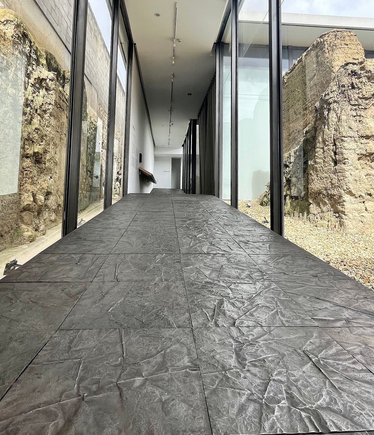 A corridor floored with large hammered grey metal tiles, glass walls on both sides revealing preserved stone ruins.
